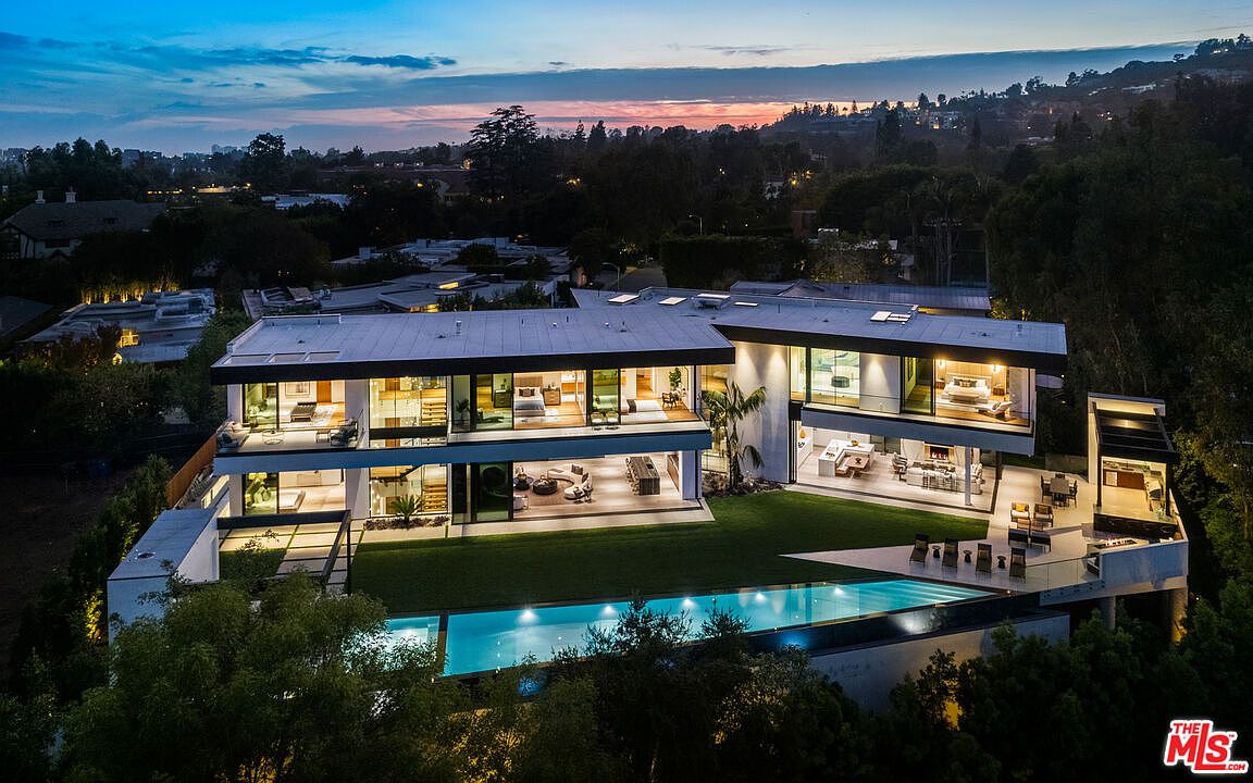 This aerial view showcases a modern, multi-level home with expansive glass walls, a flat roof, and a sleek design. A long, rectangular pool is a prominent feature, surrounded by manicured landscaping and mature trees. The property exudes luxury and privacy, set against a backdrop of distant city lights and a twilight sky.