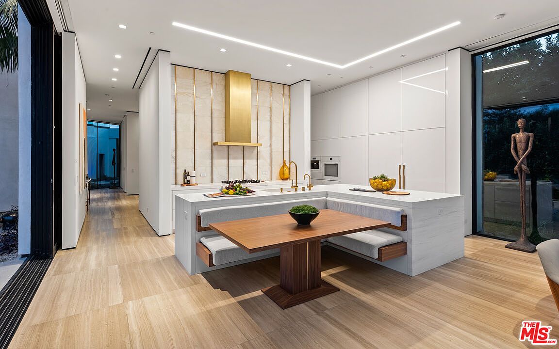 This is a modern kitchen featuring a unique built-in dining area with banquette seating. The kitchen boasts sleek white cabinetry, a marble-clad island, and a striking gold-toned range hood. Large windows provide natural light and views of the outdoors, enhancing the contemporary and luxurious feel of the space.