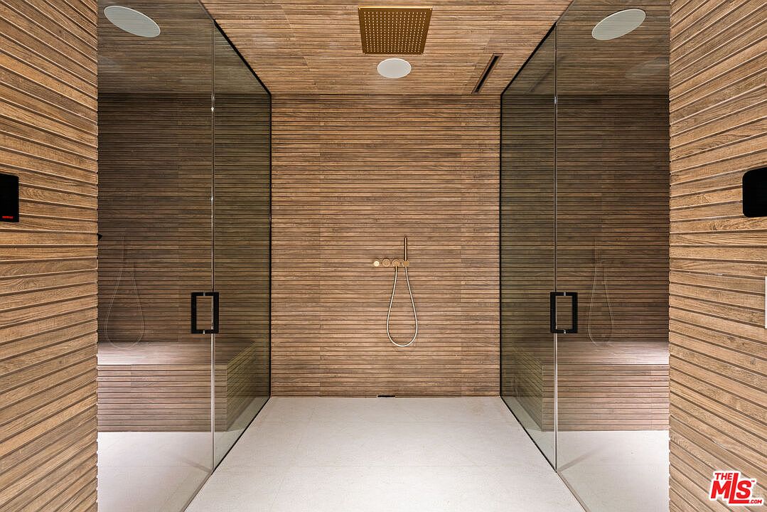 This is a luxurious primary bathroom featuring a modern, spa-like shower. The shower is enclosed with glass panels and has a built-in bench. The walls and ceiling are clad in horizontal wood paneling, and the floor is a light-colored tile. A rainfall showerhead and a handheld shower fixture are visible, suggesting a high-end experience.