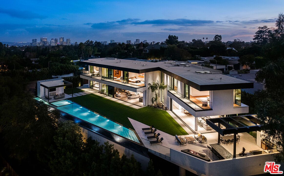 This aerial view showcases a modern, multi-level home with a sleek, flat roof and expansive glass windows. A long, narrow pool runs parallel to the house, complemented by a patio area with lounge chairs and a dining set. The property is surrounded by lush greenery, with a cityscape visible in the distance, creating a luxurious and private setting.