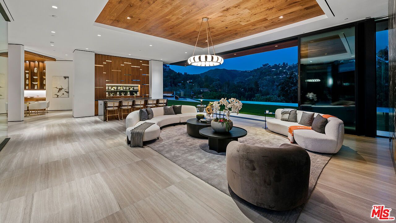 This is a luxurious living room featuring a modern design with curved sofas, round coffee tables, and a wooden ceiling. The room opens to an outdoor view of mountains and greenery through large sliding glass doors, creating a seamless indoor-outdoor living experience. A stylish bar area is visible in the background, enhancing the room's entertainment appeal.