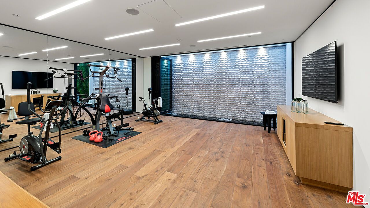 This is a spacious home gym featuring hardwood floors and a variety of exercise equipment, including stationary bikes and a weight machine. A textured wall with integrated lighting adds a modern touch, while a large mirror enhances the sense of space. A console table with decorative items sits against the wall, and a television is mounted above it.