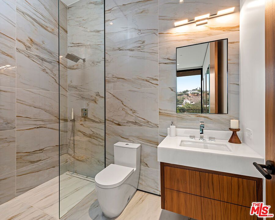 This is a modern primary bathroom featuring marble-like tile walls and flooring with gold veining. The bathroom includes a glass-enclosed shower, a toilet, and a floating vanity with a white countertop and wood-grain cabinetry. A rectangular mirror hangs above the sink, illuminated by a contemporary light fixture, creating a luxurious and clean aesthetic.