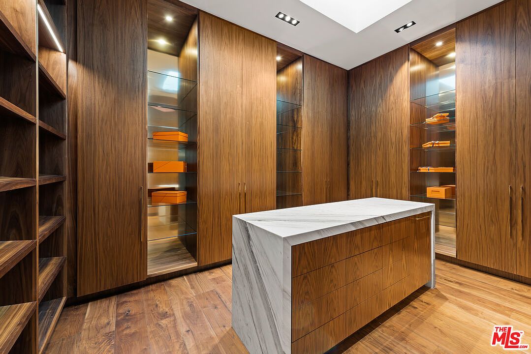 This is a luxurious walk-in closet featuring custom wood cabinetry with glass shelving and integrated lighting. A marble-topped island provides a central space for organizing accessories. The hardwood flooring and overall design create a high-end, sophisticated feel, perfect for showcasing a curated wardrobe.