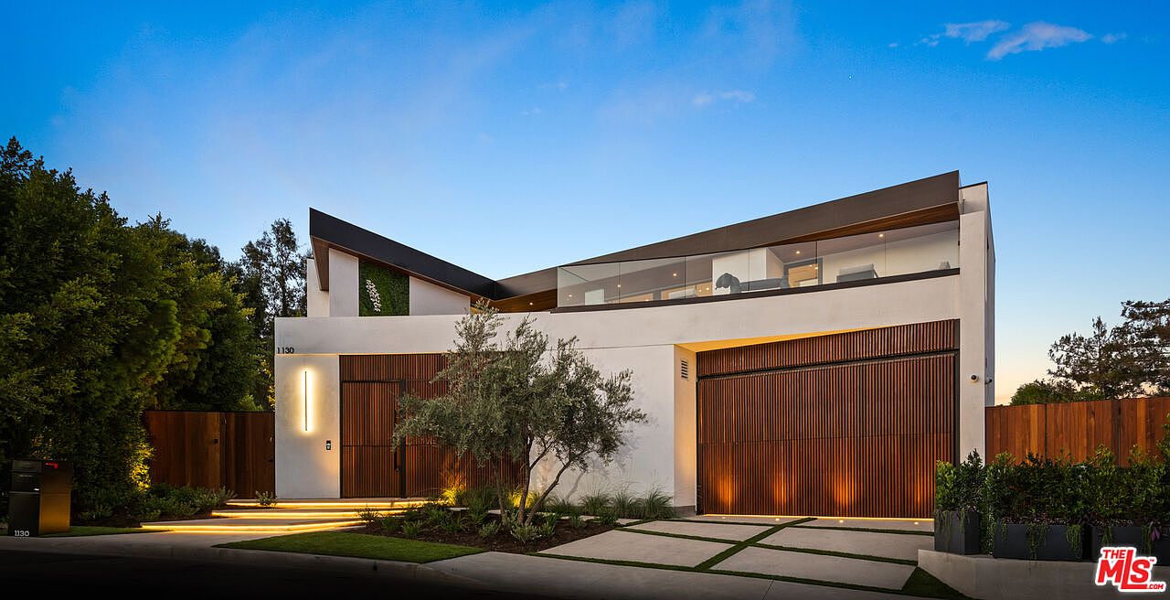 This is a front exterior view of a modern, two-story home. The house features a flat roof with an angular design, a white facade, and wooden garage doors. The landscaping includes a manicured lawn, trees, and decorative plants, enhancing the property's curb appeal.