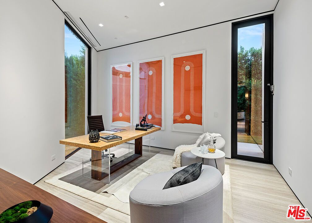 This is a modern office space featuring a light wood desk with clear acrylic legs, complemented by a brown leather office chair. The room is decorated with three large, framed orange abstract art pieces and a seating area with a gray ottoman and a small round table. Natural light floods the room through a large window and a glass door, enhancing the clean, minimalist aesthetic.