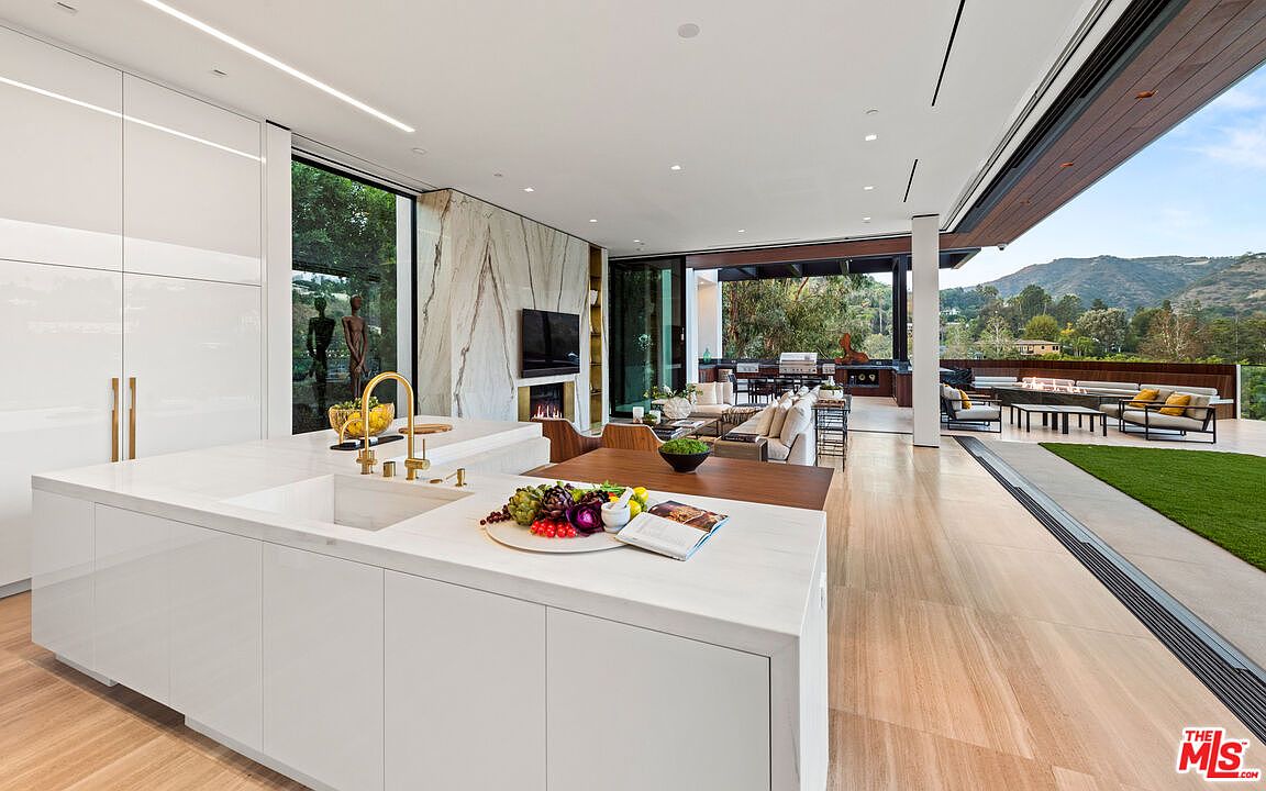 This image showcases a modern, open-concept kitchen that seamlessly blends indoor and outdoor living. The kitchen features sleek white cabinetry, a large island with a marble countertop, and high-end fixtures. Expansive sliding glass doors open to an outdoor patio with lounge seating and a view of the surrounding landscape, creating a luxurious and inviting atmosphere.
