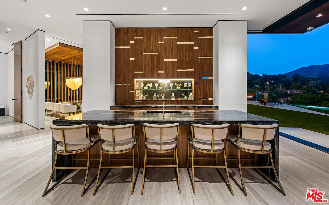 This interior shot showcases a modern kitchen bar area with five stylish bar stools featuring light-colored upholstery and gold frames. The bar top is a dark, marbled stone, complemented by a wooden backdrop with gold accents and a display case filled with glassware. Large windows offer a view of the exterior landscape, blending indoor and outdoor living.