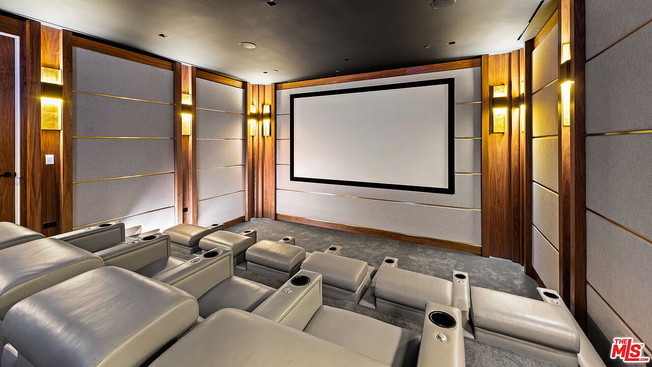 This is a luxurious home theater featuring tiered seating with plush leather recliners and integrated cup holders. The walls are adorned with sound-dampening panels accented by elegant wood trim and sconce lighting, creating a sophisticated and immersive viewing experience. A large screen dominates the front of the room, promising a cinematic escape.