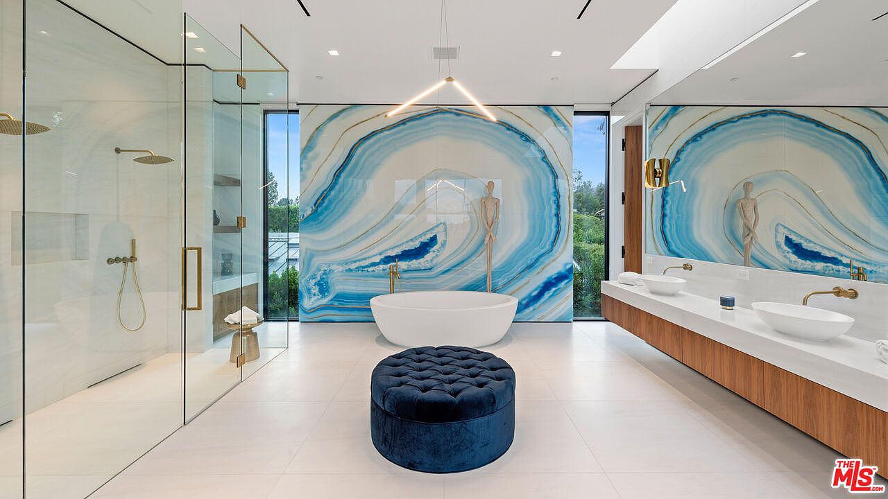 This is a luxurious primary bathroom featuring a freestanding white bathtub centered against a stunning blue and white agate-inspired wall. The room includes a glass-enclosed shower, a long floating vanity with dual sinks, and a plush blue ottoman. The design is modern and elegant, creating a spa-like atmosphere.