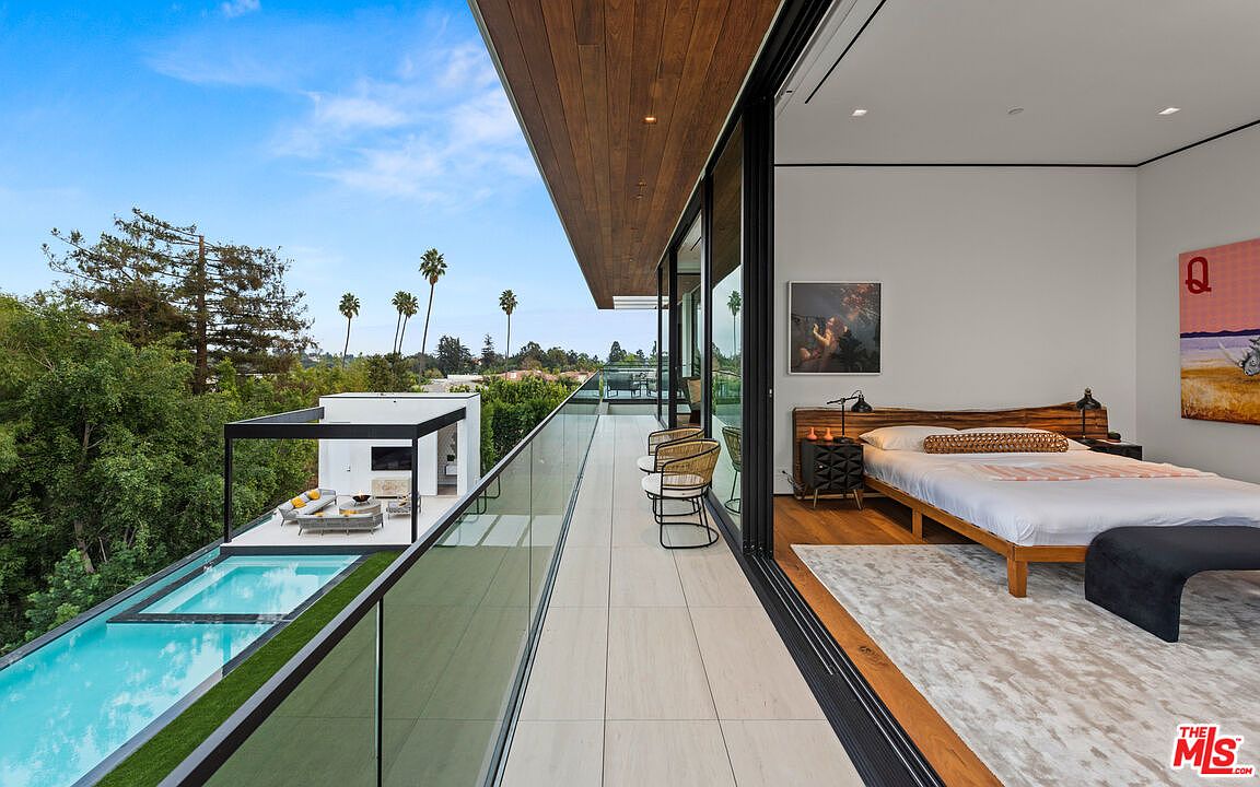 This image showcases a luxurious balcony extending from a modern bedroom, featuring sleek glass railings and stylish outdoor seating. A unique infinity pool with an adjacent lounge area is visible, offering stunning views of the surrounding landscape with palm trees and lush greenery. The open layout seamlessly connects indoor and outdoor living spaces, highlighting the property's high-end design and resort-like atmosphere.