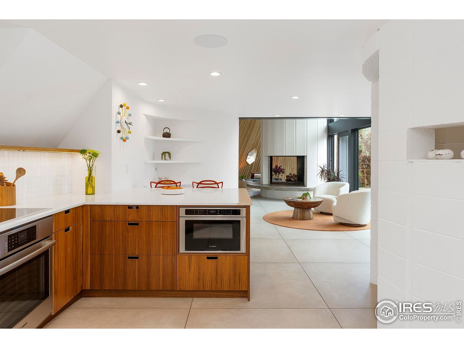 This interior shot showcases a modern kitchen that seamlessly transitions into a living area. The kitchen features sleek wooden cabinetry, stainless steel appliances, and a white countertop, while the living area boasts a fireplace, comfortable seating, and large windows that bring in natural light. The open-concept design creates a spacious and inviting atmosphere.