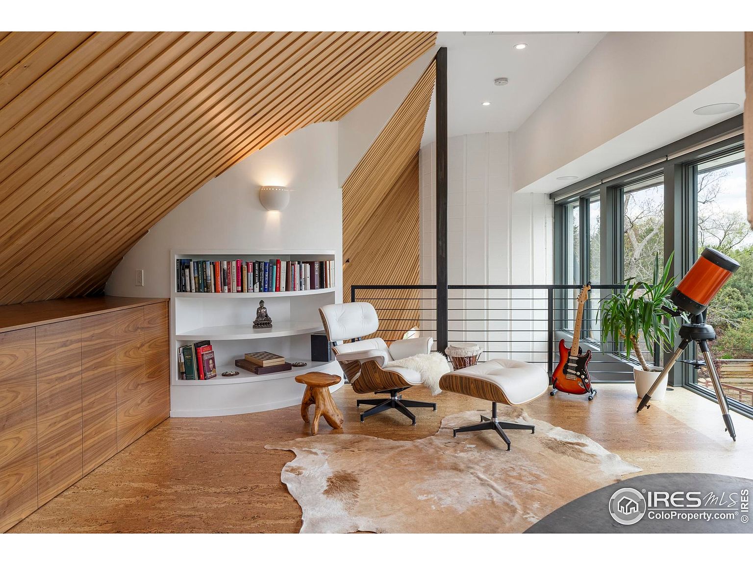 This is an interior shot of a modern living room featuring a unique architectural design with sloped wooden ceilings and a built-in bookshelf. The room is furnished with a white leather lounge chair and ottoman, a cowhide rug, and a telescope near the large windows, suggesting a space for relaxation and hobbies. The overall impression is stylish and inviting, with a focus on natural light and distinctive design elements.