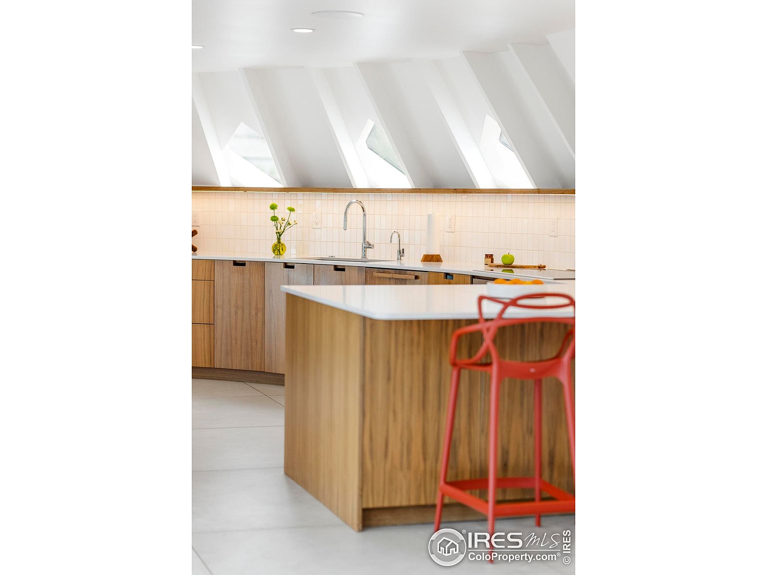 This is a bright and modern kitchen featuring light wood cabinetry and a white countertop island. The kitchen has a clean and minimalist design, with a focus on natural light from the unique angled windows. A vibrant red bar stool adds a pop of color to the space.