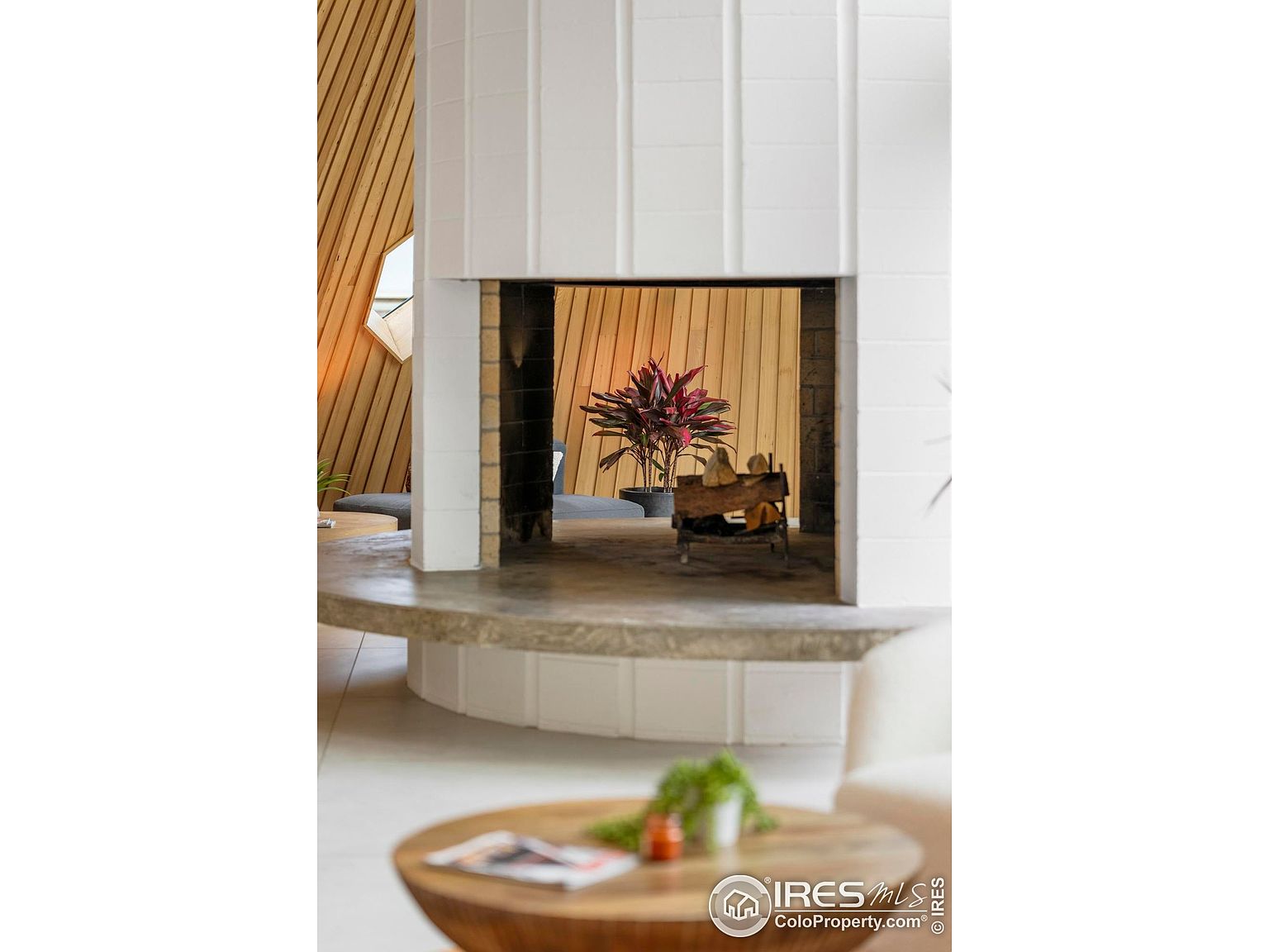 This interior shot showcases a unique fireplace design with a white tiled exterior and a view through the firebox to a wood-paneled wall and a potted plant. The fireplace is elevated on a concrete base, and a round wooden coffee table with a plant is visible in the foreground, creating a mid-century modern aesthetic. The perspective is from a low angle, emphasizing the architectural details of the fireplace.