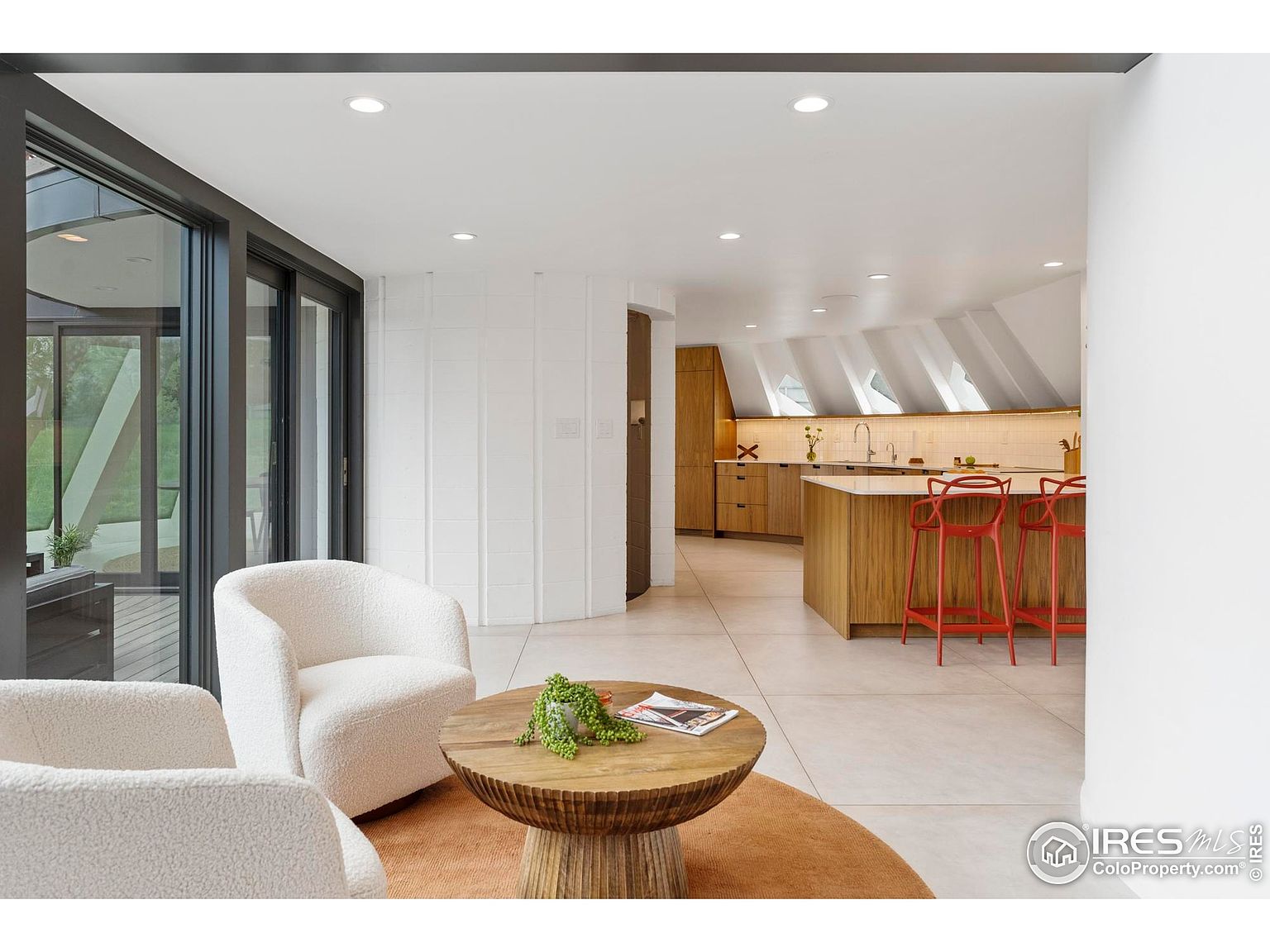 This interior shot showcases a modern living space that seamlessly blends into a kitchen area. The living room features two plush, white armchairs arranged around a unique wooden coffee table, set upon a round rug. The kitchen boasts a contemporary design with light wood cabinetry, a central island with red bar stools, and an interesting angled ceiling with skylights, creating a bright and airy atmosphere.