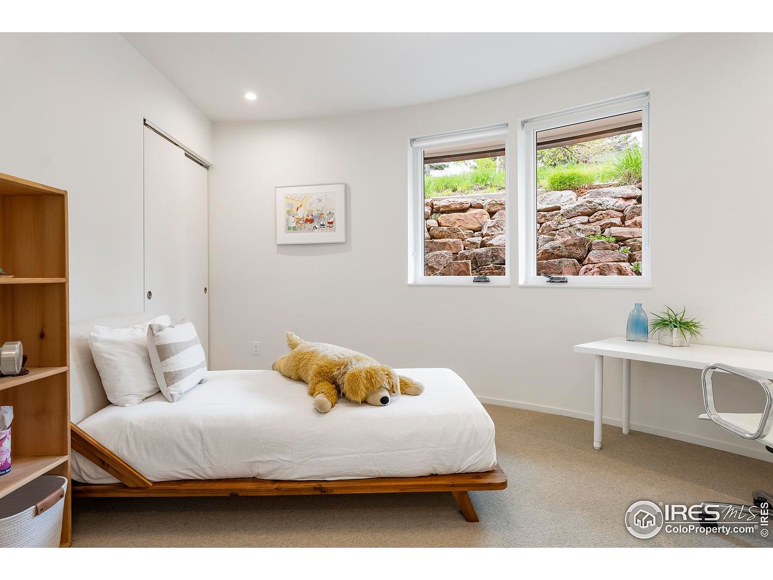 This is a bright and minimalist bedroom featuring a low-profile wooden bed frame with white linens and decorative pillows. A large stuffed dog is lying on the bed. The room has two windows offering natural light and a view of a rock wall outside, complemented by a simple white desk and chair. A wooden bookshelf is on the left side of the bed.