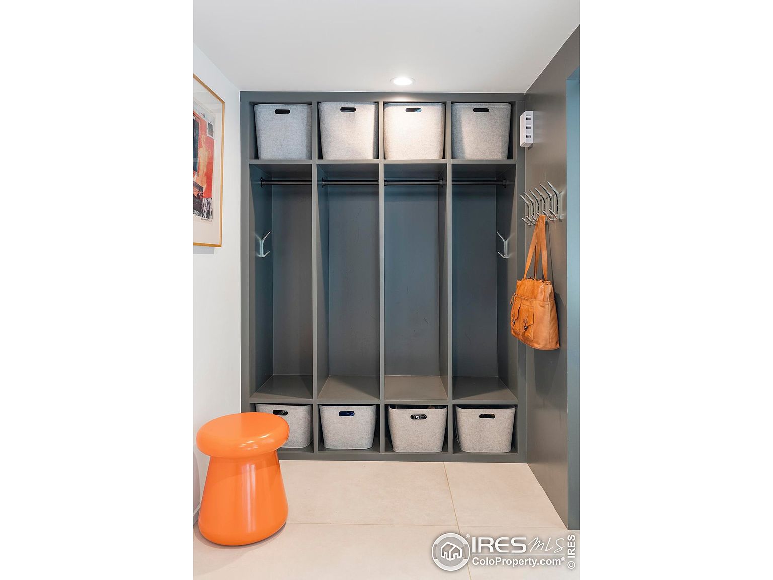 This interior shot showcases a well-organized mudroom or entryway featuring a custom-built gray storage unit with open shelving and gray storage bins. An orange stool adds a pop of color, and hooks on the side provide space for hanging items. The overall impression is clean, modern, and functional, ideal for a family home.
