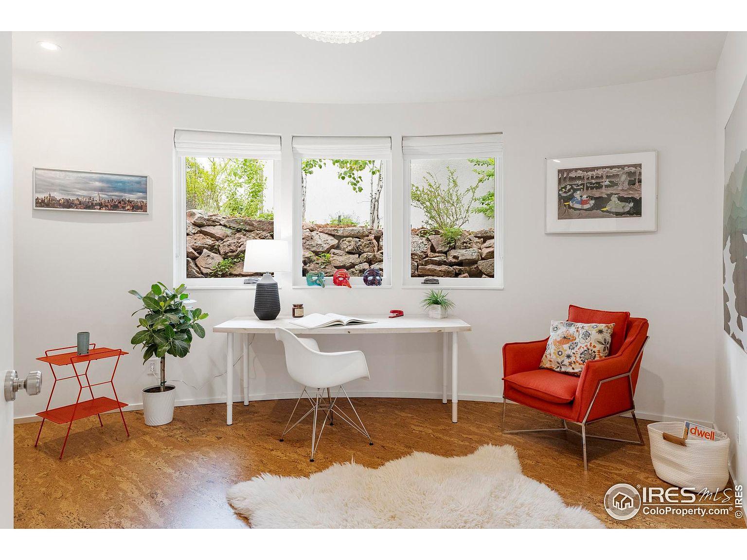 This is an interior shot of a bright and airy home office. The room features a white desk, a modern white chair, and an orange armchair, complemented by a white faux fur rug. Three windows offer natural light and a view of an exterior rock wall and greenery, enhancing the room's serene atmosphere.