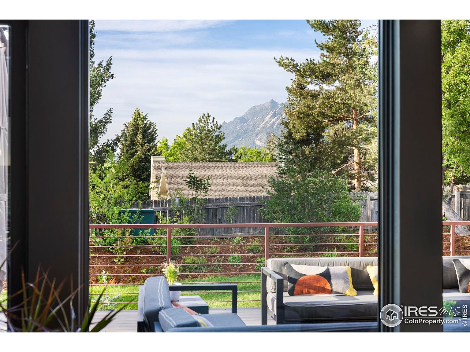 This image showcases a stylish outdoor patio or deck area, framed by a modern doorway. The space features comfortable seating arrangements with contemporary cushions, set against a backdrop of lush greenery and a mountain view. A wooden fence and railing add to the privacy and charm of the outdoor living space.