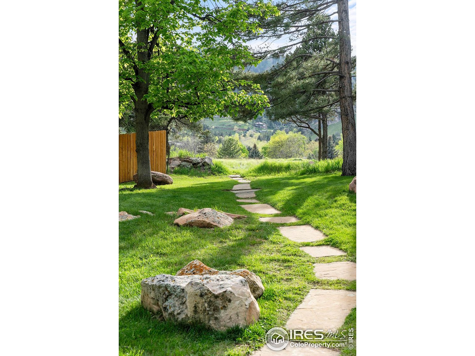 This image showcases a beautifully landscaped yard or garden, featuring a stone pathway that meanders through lush green grass. Large rocks are strategically placed throughout the yard, adding a natural and rustic touch. The scene is framed by mature trees, creating a serene and inviting outdoor space.