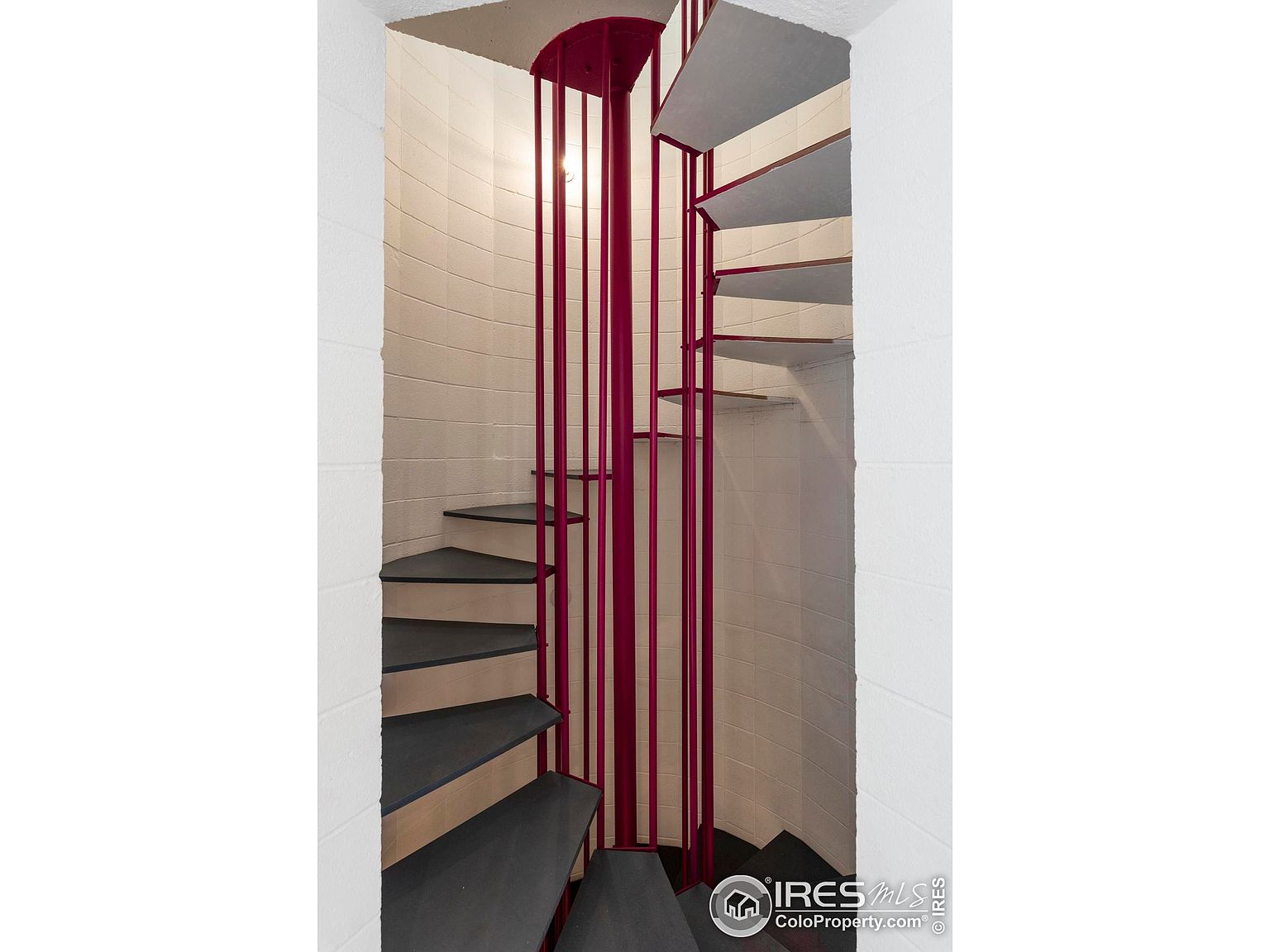 This interior shot showcases a unique spiral staircase with a striking red support structure. The stairs themselves are a dark gray, contrasting with the light-colored, textured walls of the stairwell. The design creates a visually interesting and space-saving solution for accessing different levels of the home.