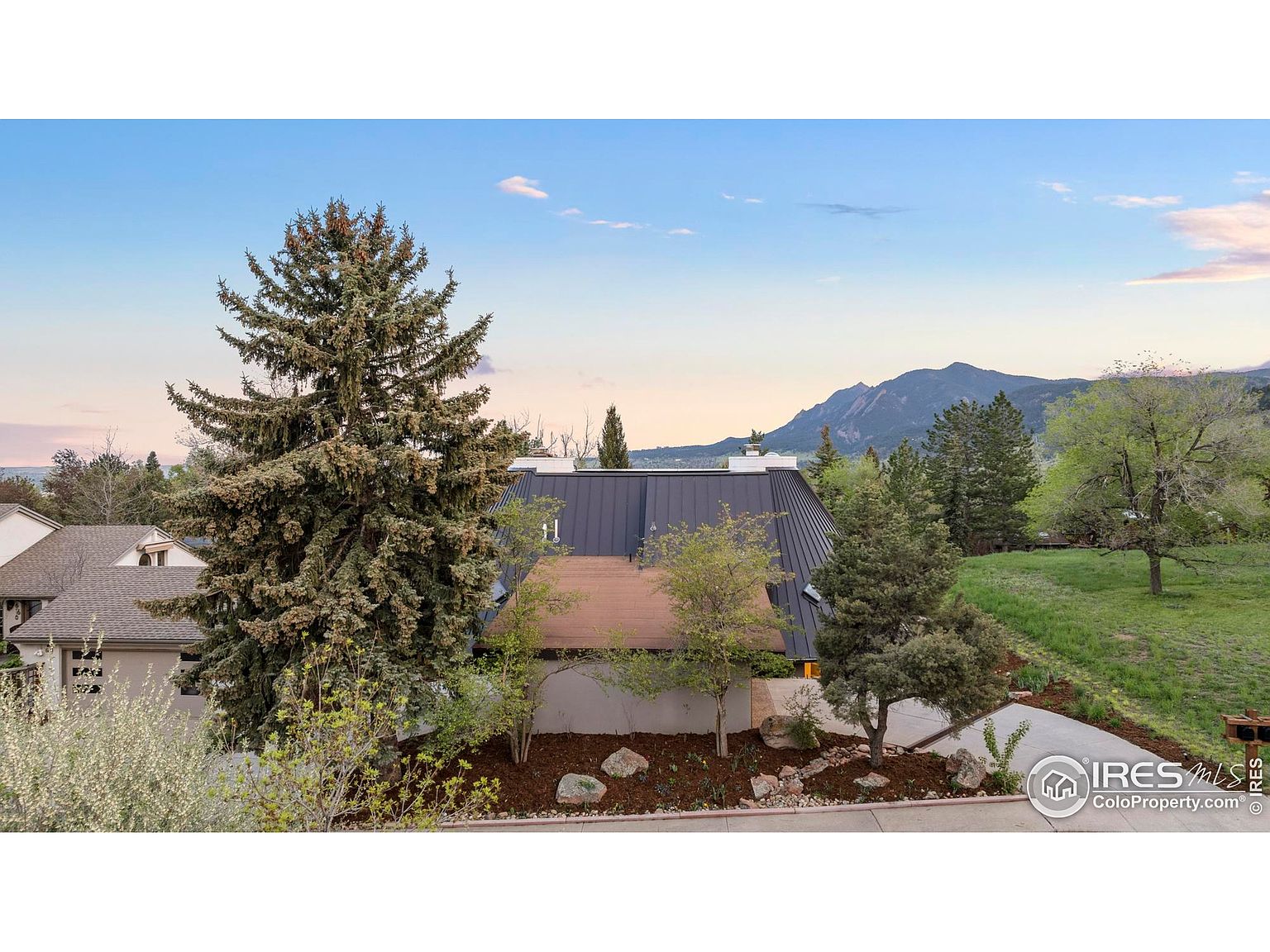 This is a front exterior view of a house with a dark metal roof and a mix of natural landscaping. A large evergreen tree dominates the left side of the frame, while smaller trees and shrubs adorn the front yard. The house is set against a backdrop of mountains and a blue sky, creating a serene and inviting atmosphere.