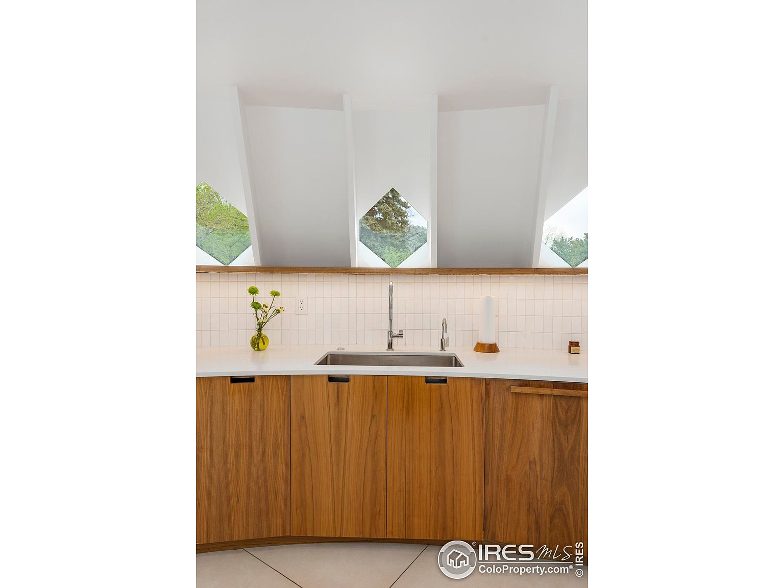 This is a well-lit kitchen featuring wooden cabinets with sleek, minimalist hardware. A stainless steel sink is set into a white countertop, and a tiled backsplash adds texture. Unique diamond-shaped windows above the cabinets provide natural light and a view of the outdoors.