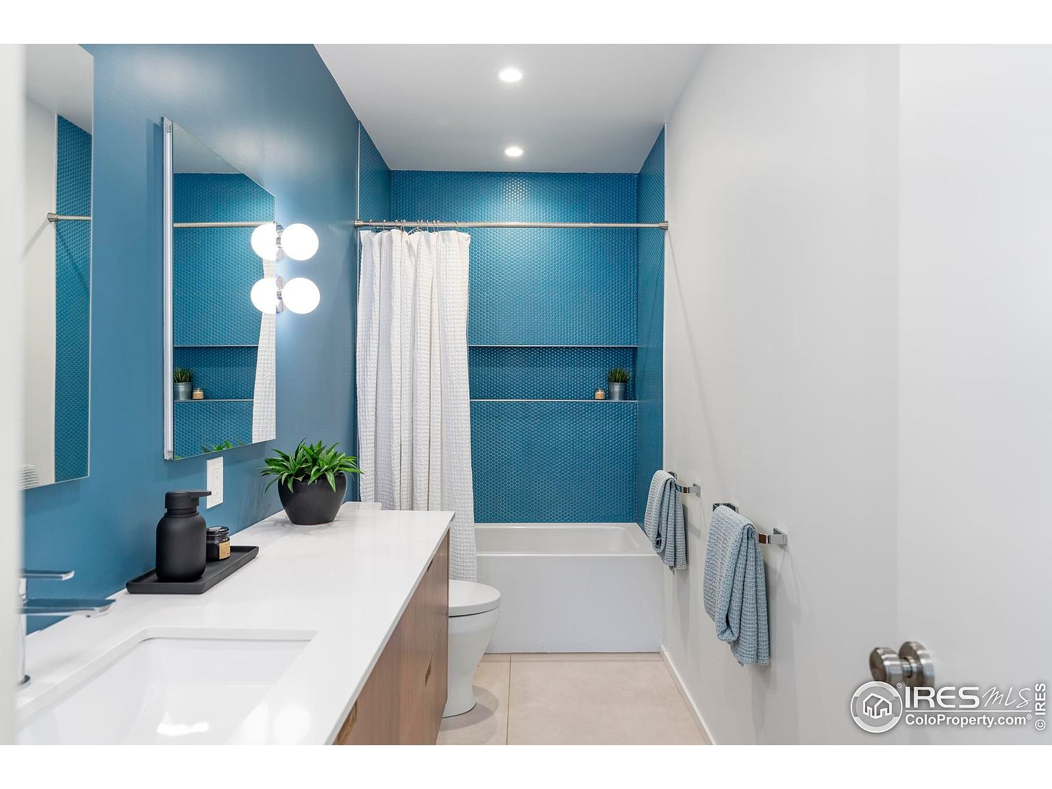 This is a well-lit bathroom featuring a modern design. The walls are painted in a combination of blue and white, with blue honeycomb tiles accenting the shower area. A white countertop sits above a wooden vanity, complemented by a sleek sink and modern faucet. The bathroom includes a bathtub with a shower curtain, a toilet, and towel racks with neatly folded towels.