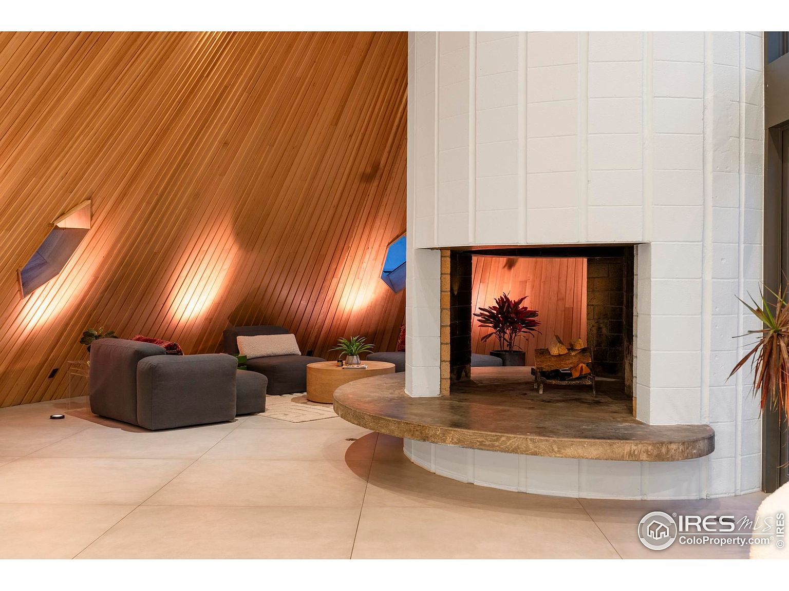 This interior shot showcases a unique living room with a striking architectural design. The room features a prominent white brick fireplace, complemented by warm wood paneling on the walls. Comfortable gray seating is arranged around a circular coffee table, creating an inviting and stylish space.
