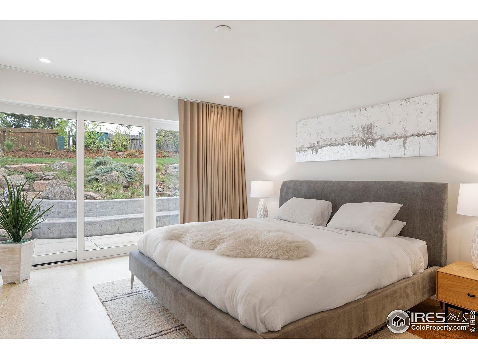 This is a bright and airy primary bedroom featuring a large bed with a gray upholstered frame and a white comforter. A sliding glass door provides access to the outdoor area, and neutral-toned curtains add a touch of elegance. The room is decorated with a minimalist aesthetic, including a piece of artwork above the bed and a potted plant near the door.