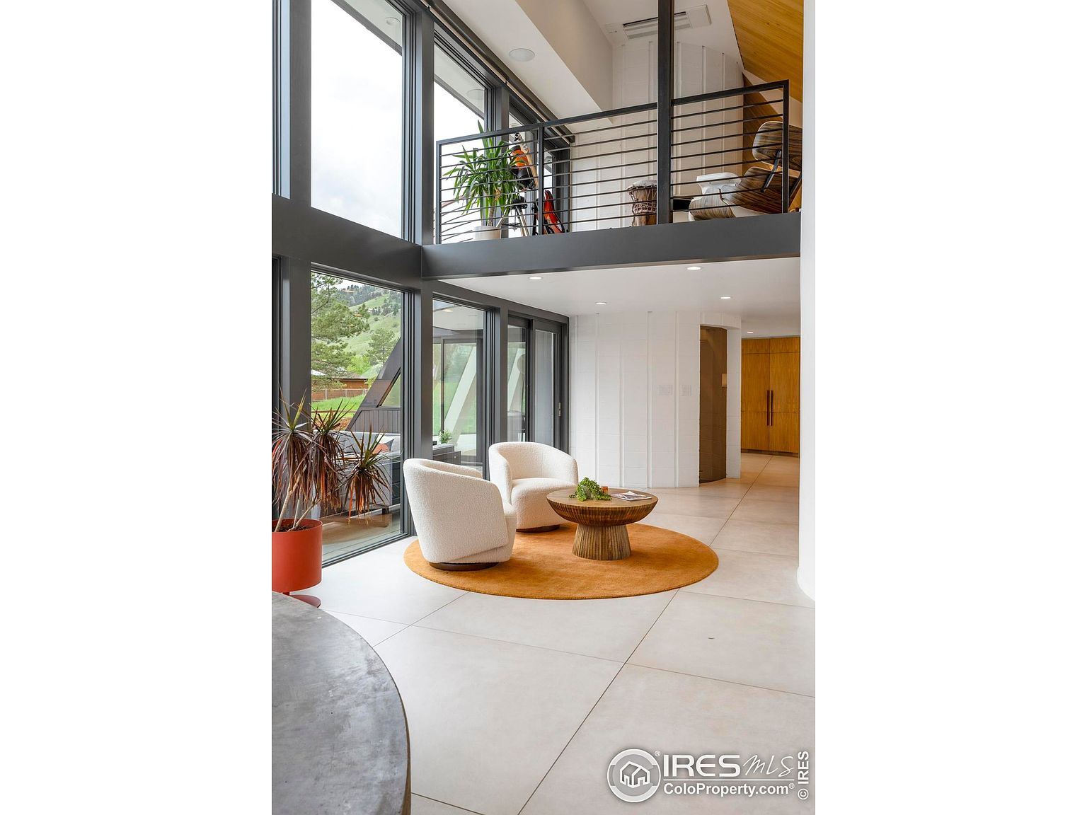 This interior shot showcases a modern living room with a double-height ceiling and large windows offering natural light and views of the outdoors. The room features two armchairs on a round rug, a unique coffee table, and a balcony overlooking the space. The overall design is contemporary and airy, emphasizing the connection between indoor and outdoor living.