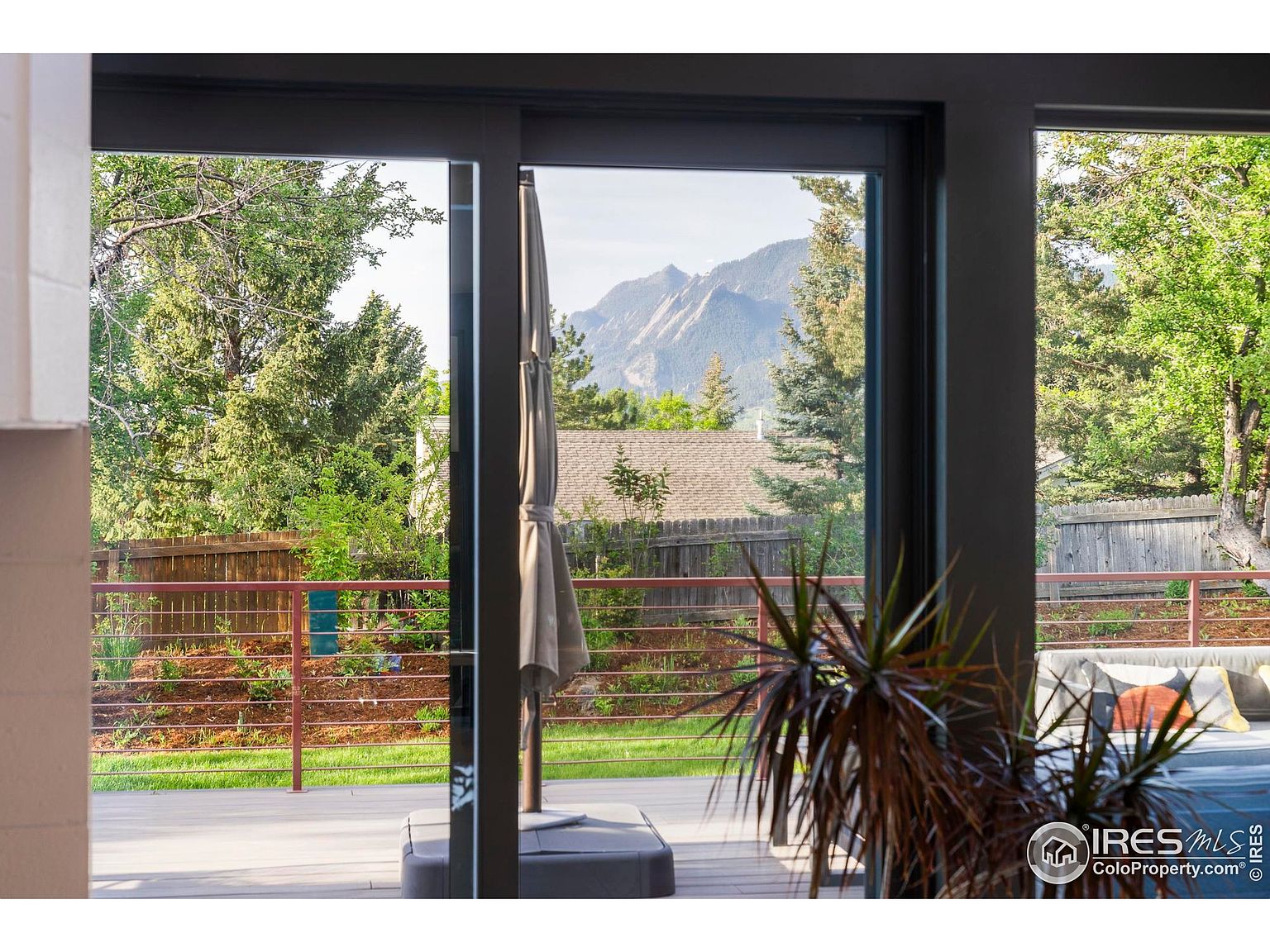 This image showcases a modern patio or deck area, viewed through a large sliding glass door. The deck features a sleek railing system and outdoor furniture, suggesting a comfortable space for relaxation and entertainment. In the background, there are trees and a mountain view, enhancing the appeal of the outdoor living area.