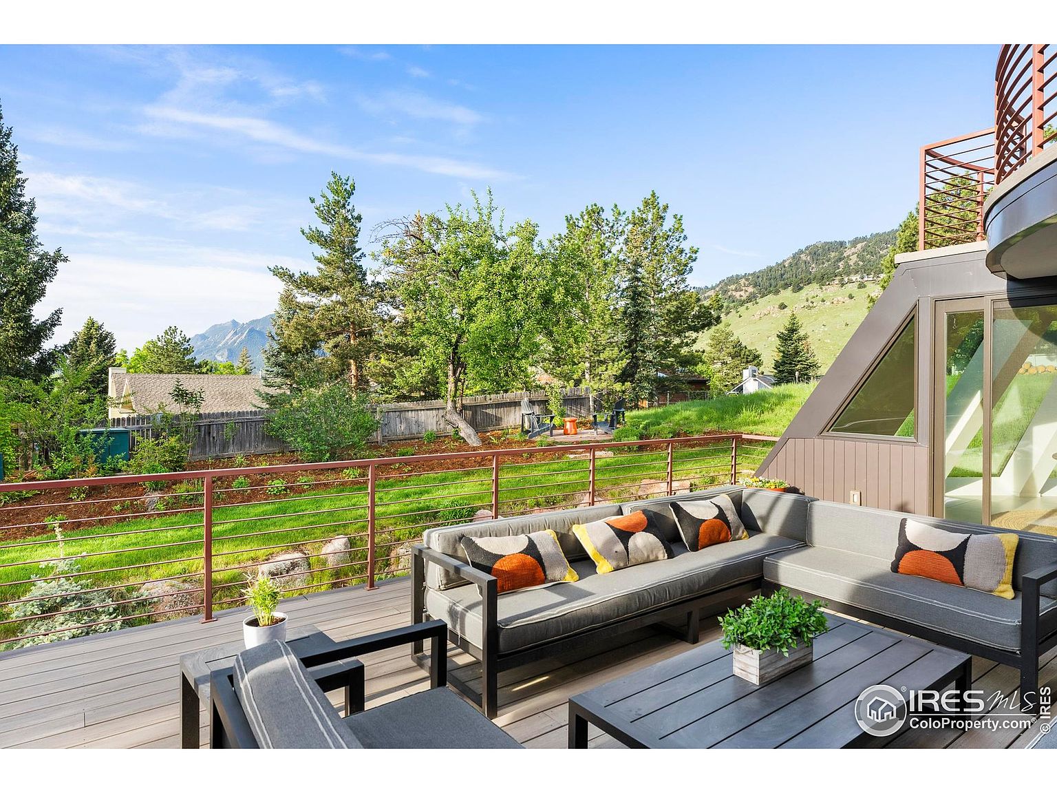 This image showcases a beautifully designed outdoor living space, featuring a spacious deck with comfortable gray seating and stylish accent pillows. The deck overlooks a lush green lawn and garden area, with a mountain view in the background. The space is perfect for relaxation and entertaining, offering a seamless blend of indoor and outdoor living.