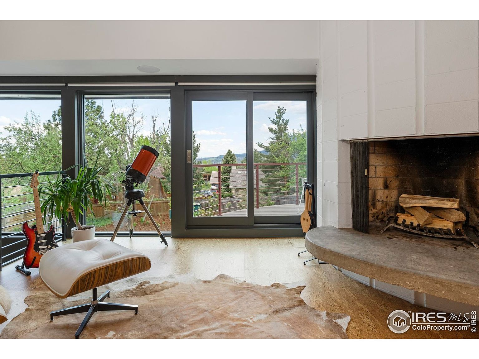 This is an interior shot of a living room featuring a modern design with large windows offering a view of the outdoors. The room includes a fireplace, a cowhide rug, a lounge chair, a telescope, and musical instruments, creating a unique and inviting space. The natural light and open layout enhance the room's appeal.
