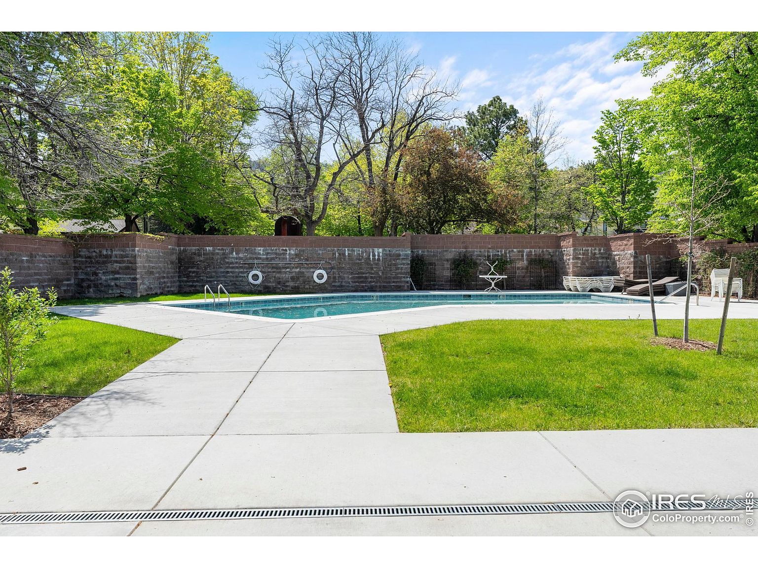 This image showcases a well-maintained outdoor swimming pool area. The pool is surrounded by a concrete patio and grassy areas, enclosed by a brick wall that provides privacy. Lounge chairs and tables are arranged around the pool, suggesting a relaxing and inviting space for recreation and leisure.