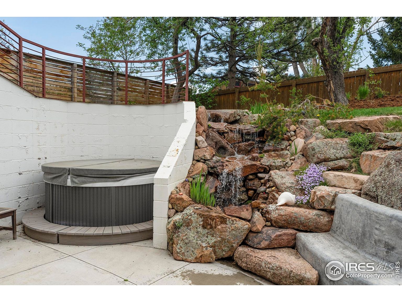 This image showcases a beautifully landscaped backyard featuring a waterfall cascading over natural rocks, creating a serene and inviting atmosphere. A hot tub is nestled against a retaining wall, offering a relaxing retreat. The garden is complemented by lush greenery and a stone bench, enhancing the overall appeal of the outdoor space.