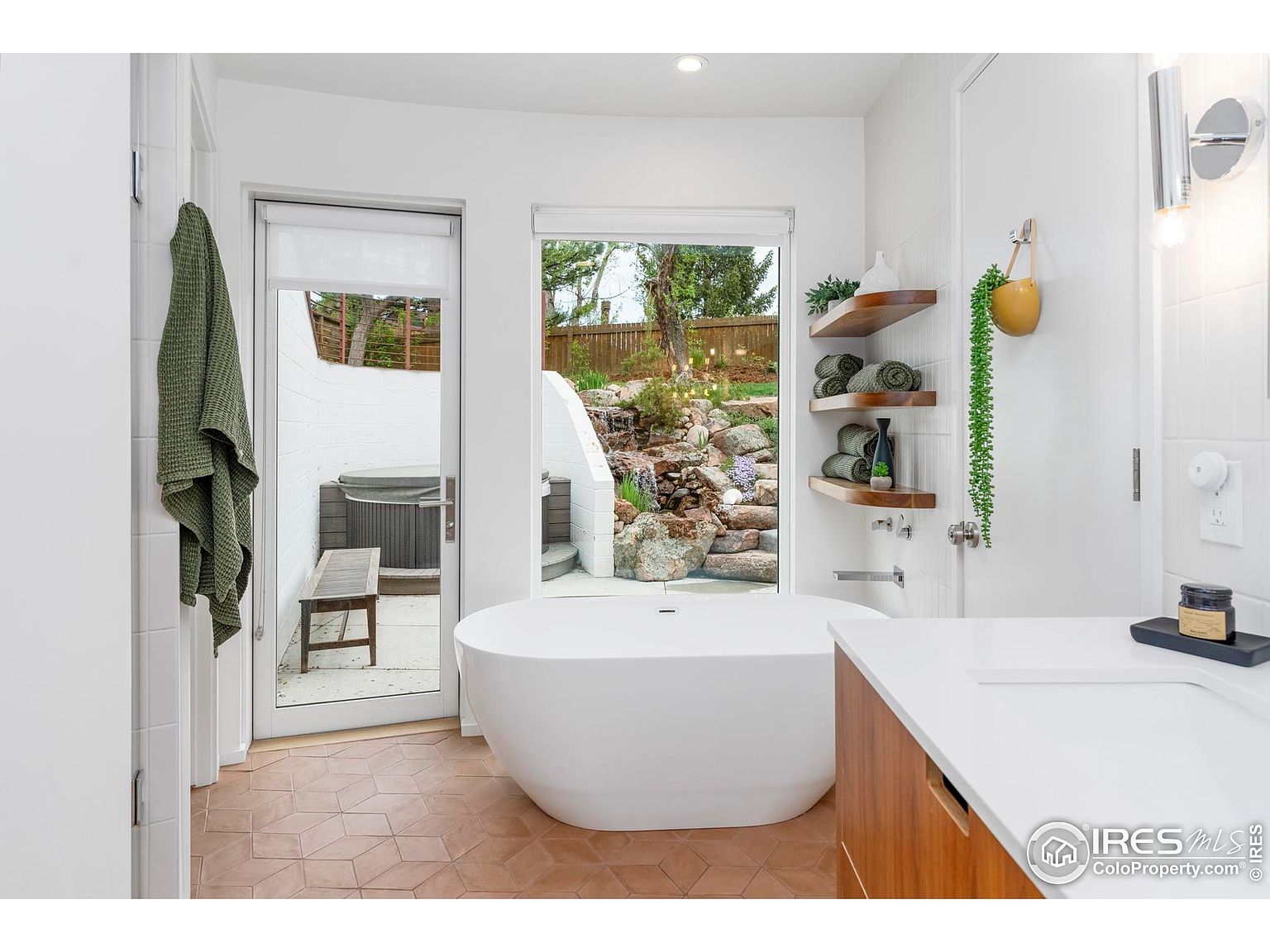 This is a bright and modern primary bathroom featuring a freestanding soaking tub positioned in front of a large window with a view of an outdoor spa. The bathroom has a clean, minimalist design with white walls, wood accents, and unique geometric floor tiles. Open shelving displays neatly rolled towels and decorative items, adding a touch of warmth to the space.