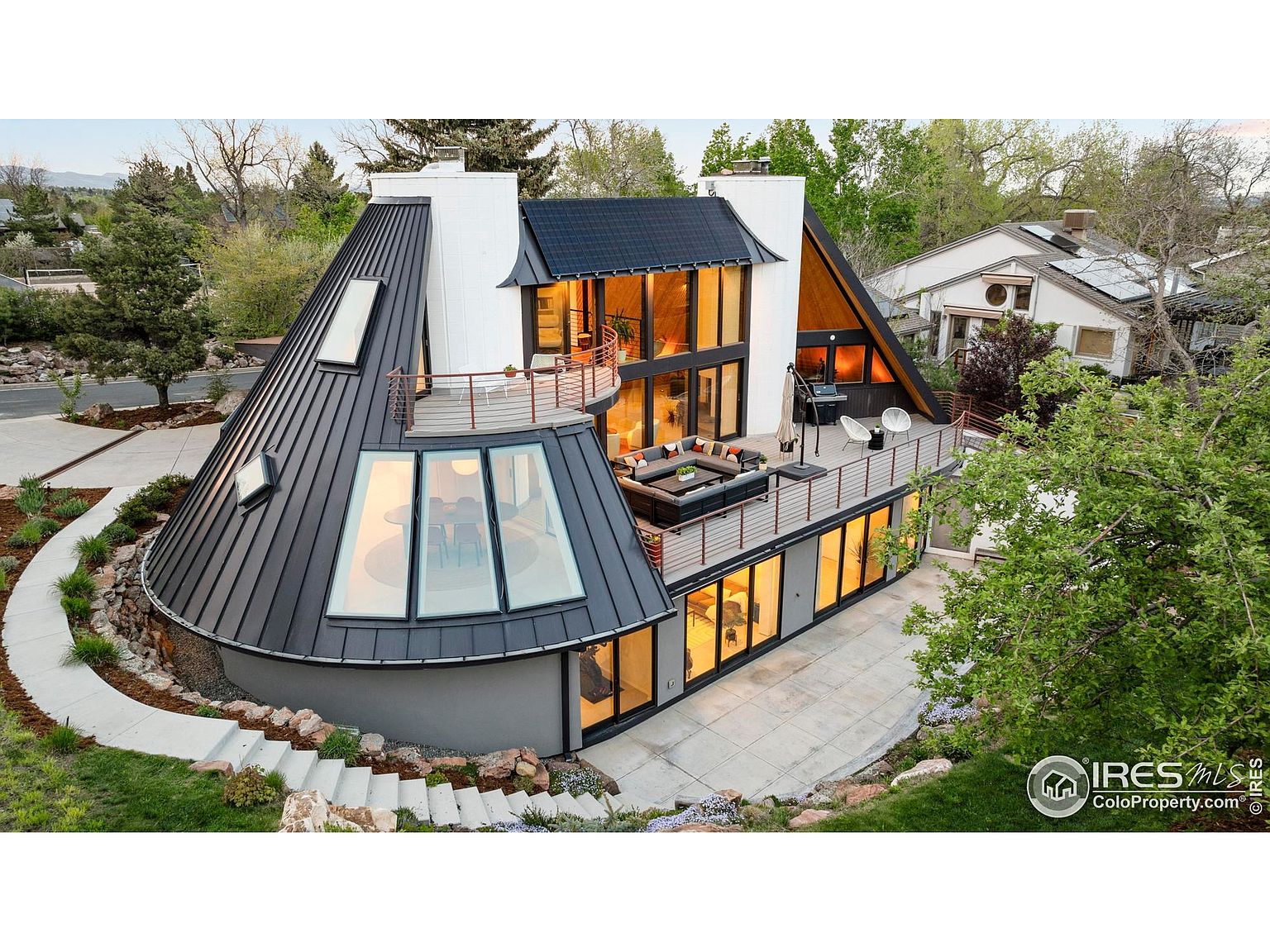 This aerial view showcases a unique, modern home with a distinctive architectural design. The property features a combination of geometric shapes, including a cone-shaped structure with skylights and a flat-roofed section with a balcony. The exterior is complemented by well-maintained landscaping and a patio area, creating an inviting and stylish living space.