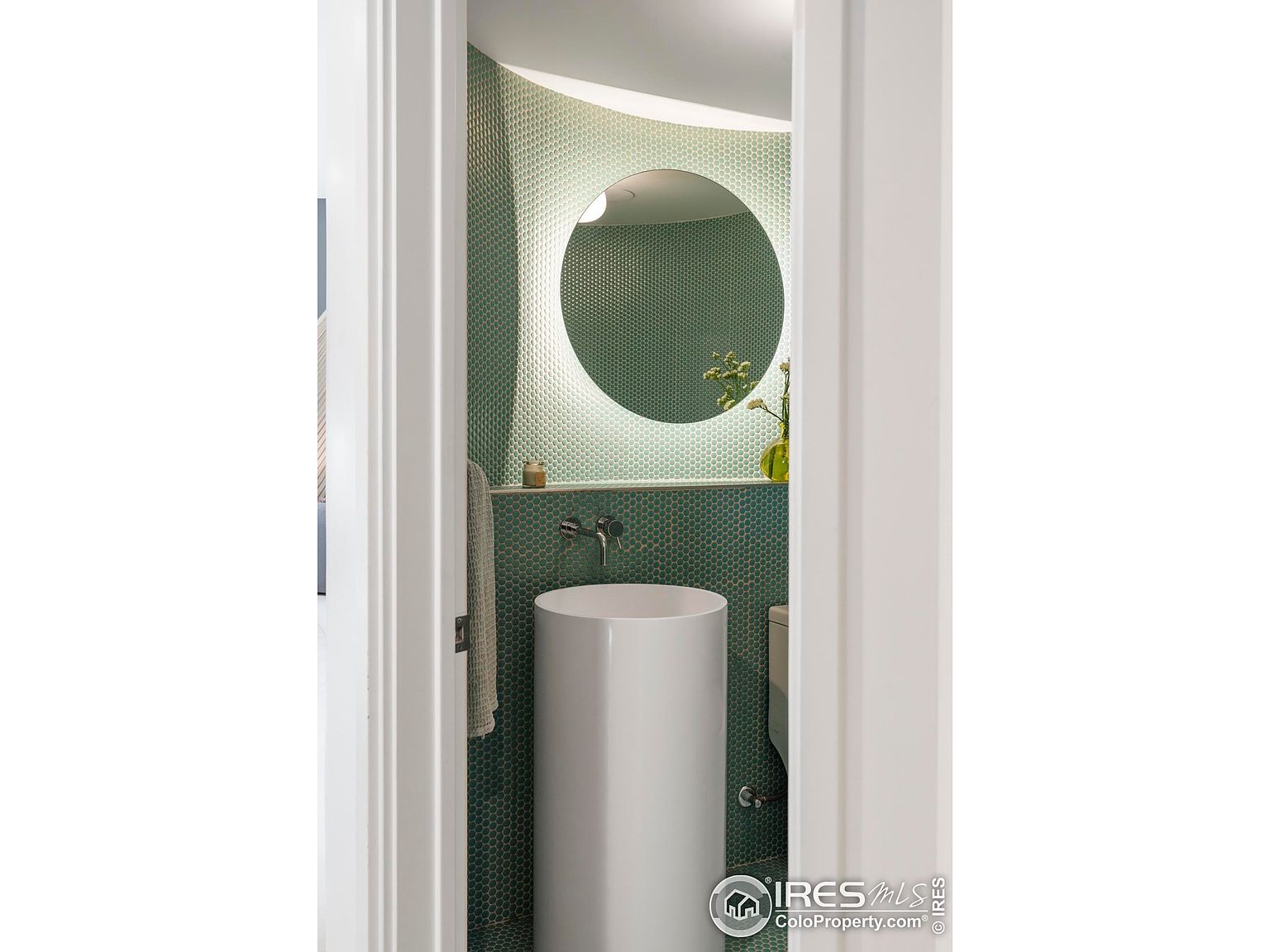 This image showcases a modern, compact bathroom with a unique design. The walls are covered in small, green hexagonal tiles, complemented by a round, illuminated mirror. A sleek, cylindrical pedestal sink stands prominently, adding to the bathroom's minimalist and stylish aesthetic.