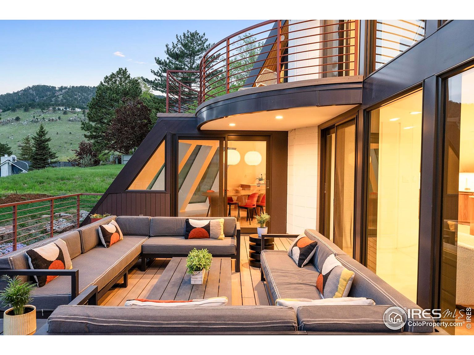 This image showcases a stylish outdoor patio/deck area, featuring a large sectional sofa with decorative pillows and a wooden coffee table. The space is part of a modern home with large glass doors and windows, offering seamless indoor-outdoor living. The deck overlooks a green lawn and distant hills, creating a serene and inviting atmosphere.