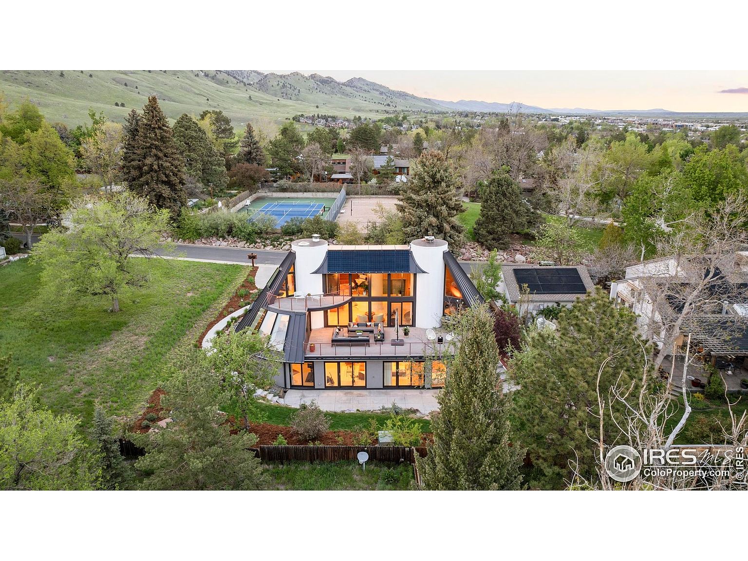 This aerial view showcases a modern, architecturally unique home nestled among lush greenery. The property features a flat roof, large windows, and cylindrical tower-like structures, offering ample natural light and views. A tennis court is visible in the background, suggesting recreational amenities, while the surrounding landscape provides privacy and a sense of tranquility.