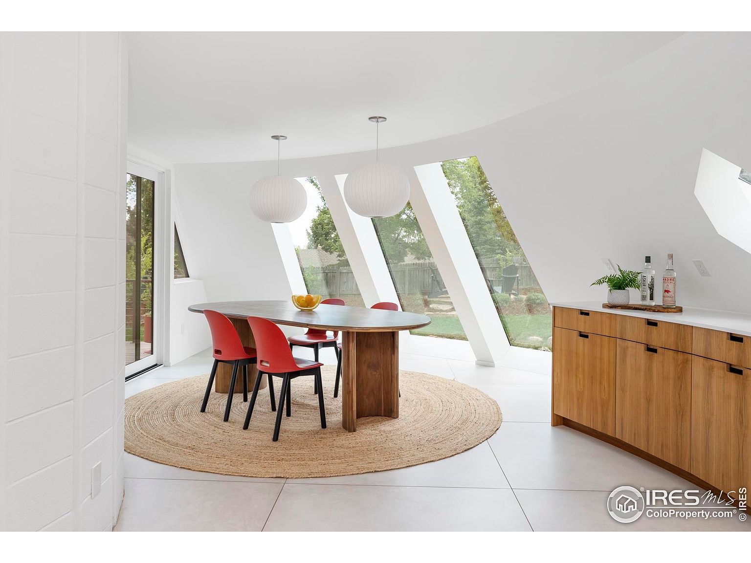 This is a bright and airy dining room featuring a unique architectural design with angled windows. The room is furnished with a wooden oval dining table surrounded by red chairs, set upon a round jute rug. A built-in wooden cabinet with a white countertop adds storage and display space, enhancing the room's modern and stylish appeal.