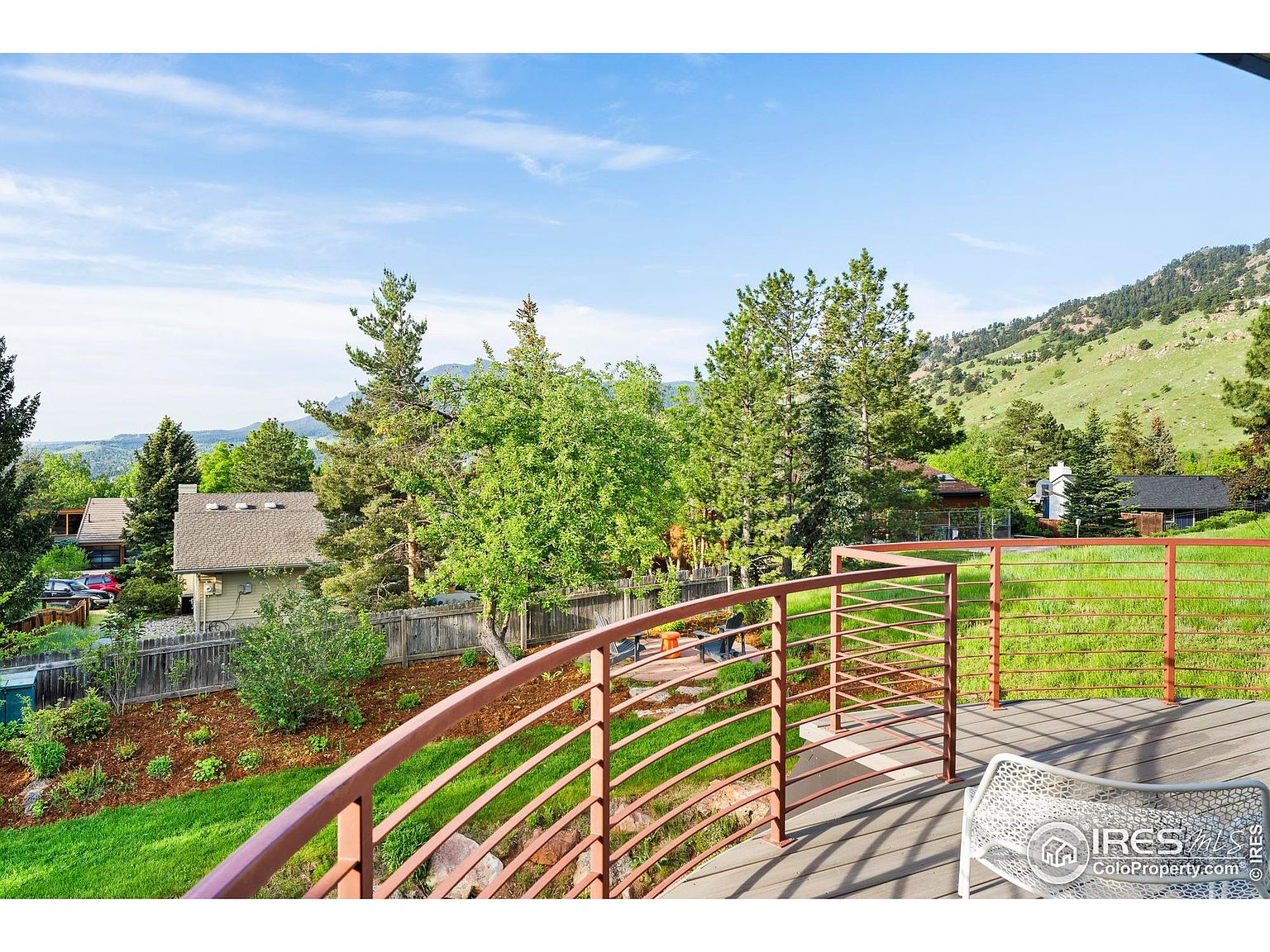 This image showcases a deck with a unique curved railing, offering a view of a lush green lawn, mature trees, and a hillside in the background. The deck features wooden flooring and a white chair, creating a relaxing outdoor space. The scene suggests a peaceful and private setting, ideal for enjoying the natural surroundings.