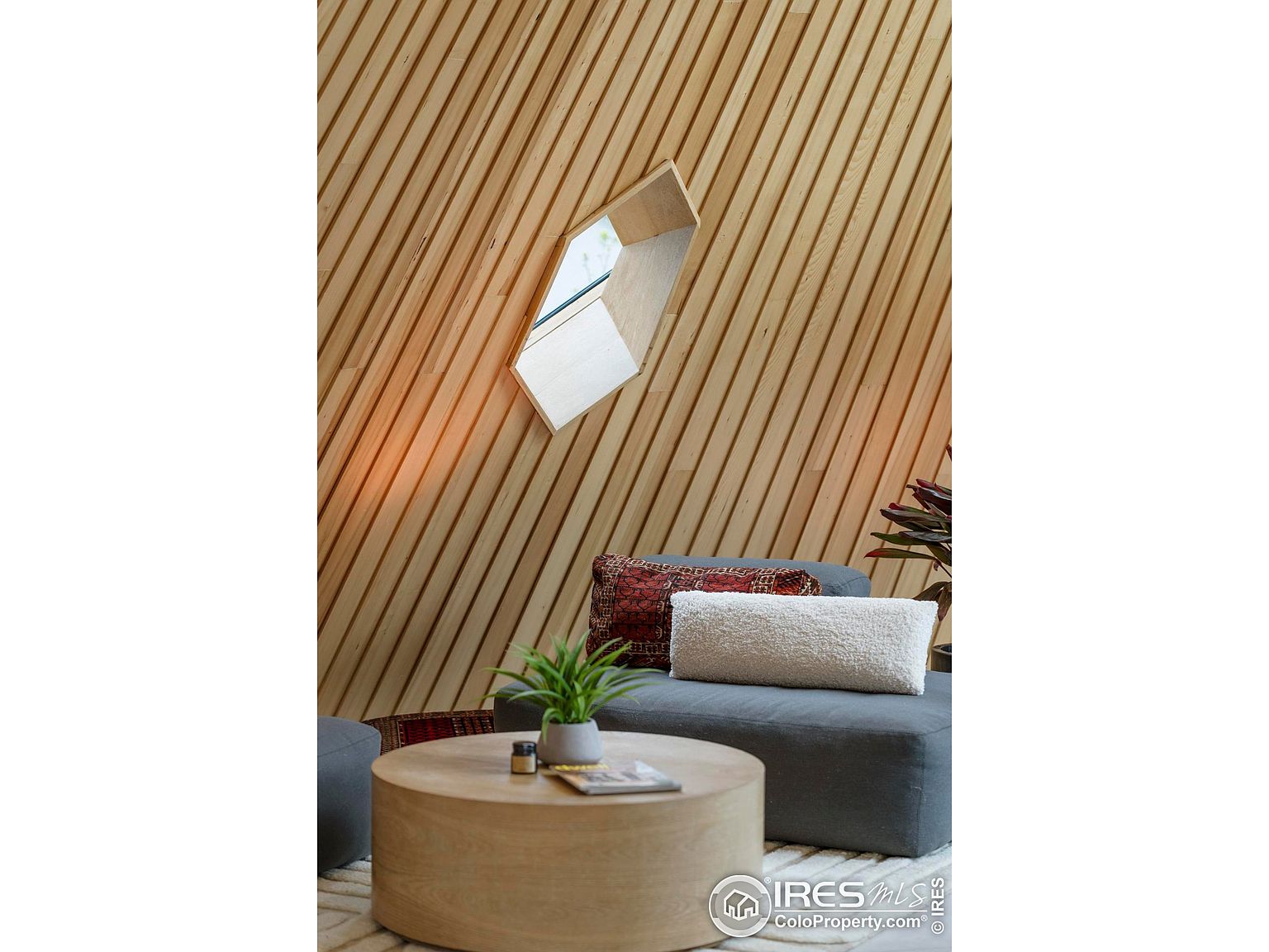 This interior shot showcases a modern living room area with a unique architectural window and wood-paneled wall. A round coffee table sits atop a light-colored rug, complemented by a gray ottoman and decorative pillows. The space exudes a minimalist yet cozy atmosphere, perfect for relaxation and conversation.