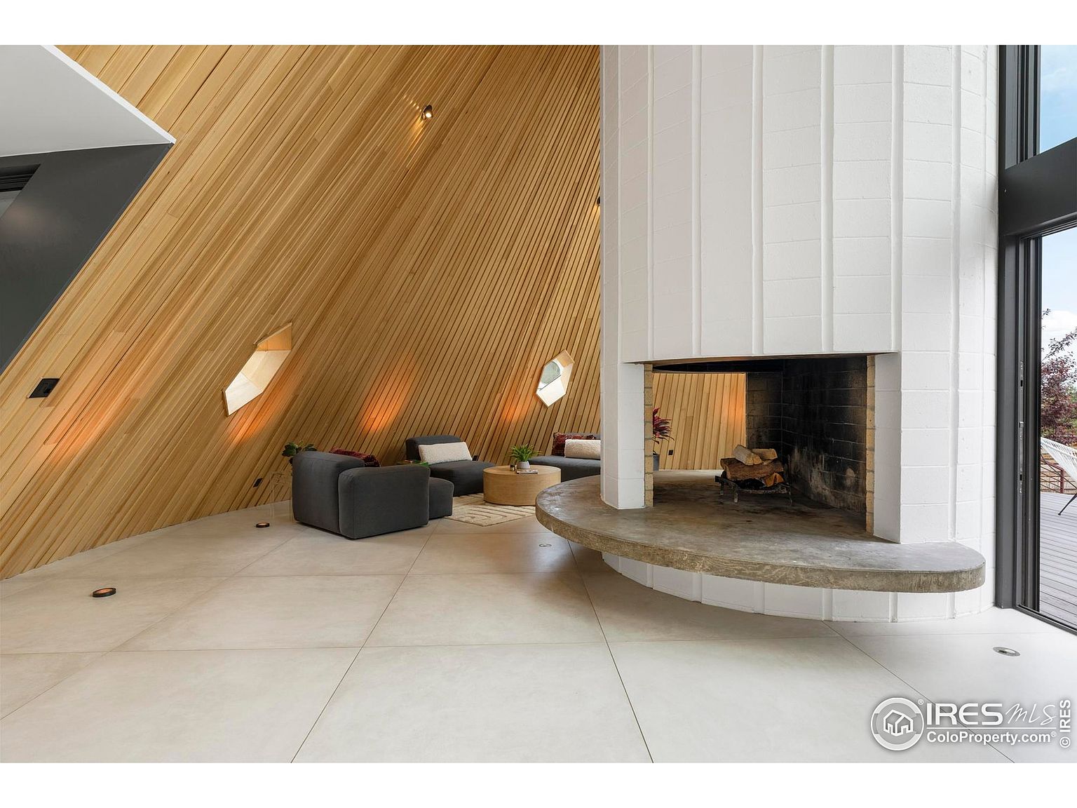 This interior shot showcases a unique living room with a striking architectural design. The room features a slanted wooden wall with small, angular windows, complemented by a modern white fireplace and neutral-toned furniture. The open space and minimalist decor create a sophisticated and inviting atmosphere.