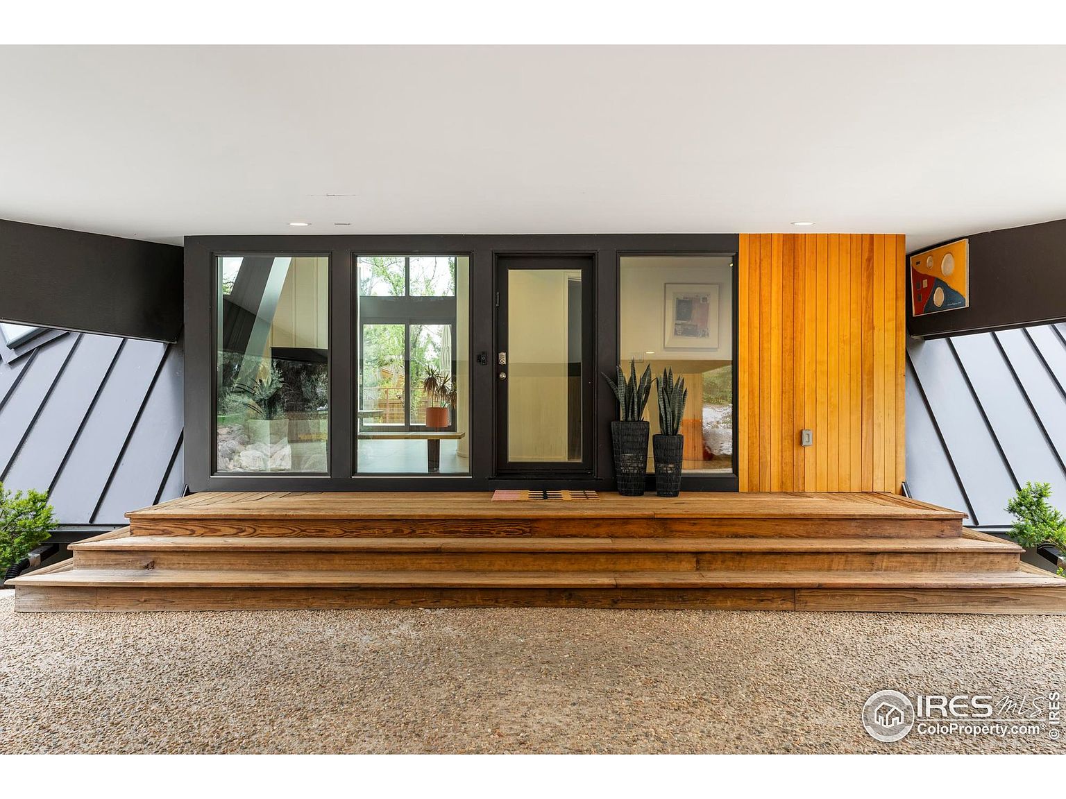 This is an inviting entryway to a modern home, featuring a wide wooden deck leading to a dark-framed glass door and windows. The exterior is accented with vertical wood paneling and a gravel ground cover. The overall impression is clean, contemporary, and welcoming.