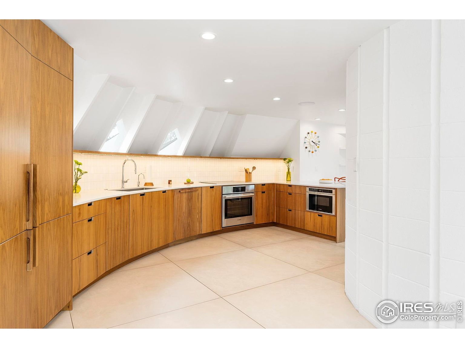This is a well-lit kitchen featuring curved wooden cabinetry and a white countertop. The kitchen has a modern design with stainless steel appliances and a unique ceiling with skylights. The flooring is light-colored tile, and the overall impression is clean and contemporary.