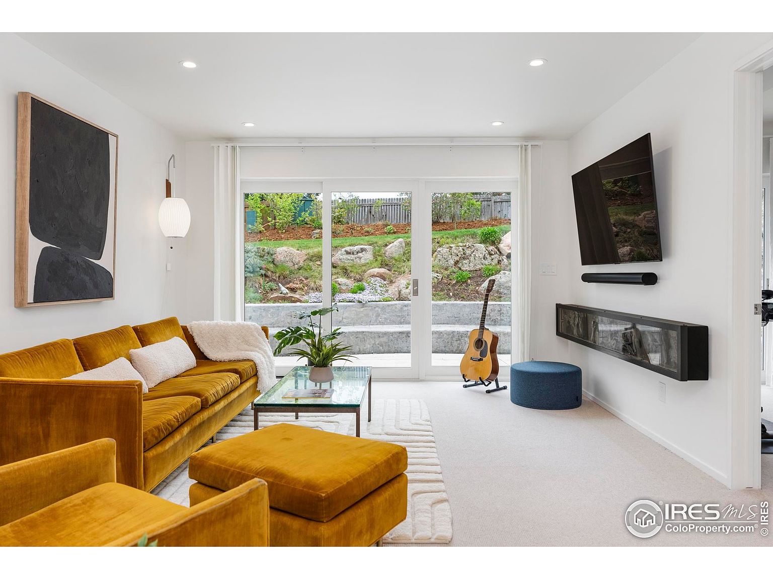 This is a bright and modern living room featuring a mustard-colored velvet sofa and ottoman, complemented by a glass coffee table and a neutral-toned rug. A large abstract artwork hangs on the wall, and a flat-screen TV is mounted above a sleek display cabinet. The room opens to an outdoor area through sliding glass doors, offering a view of a landscaped backyard.