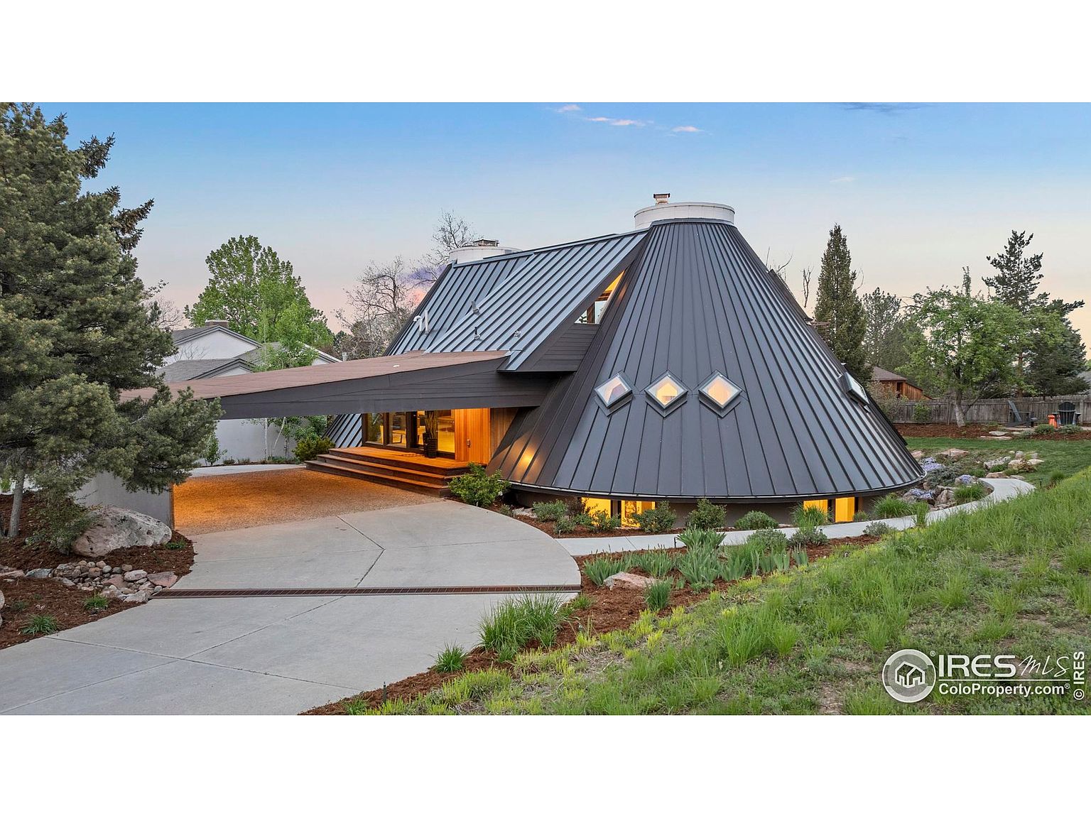 This is a striking front view of a unique, modern home featuring a conical roof design and a covered driveway. The exterior is characterized by its dark metal roofing, diamond-shaped windows, and a well-manicured lawn with landscaping. The driveway leads to a covered entrance, adding to the home's distinctive architectural appeal.